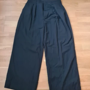 Svarta wide kostymbyxor Gina Tricot - Snygga svarta kostymbyxor från Gina Tricot i storlek 42. Byxorna har hög midja, vida ben och klassiska pressveck. De är tillverkade i ett mjukt tyg med bälteshällor och fickor både fram och bak. Perfekta för en clean och stilren look.