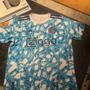Ajax blå och vit fotbollströja Adidas - Ajax fotbollströja från Adidas med blått och vitt abstrakt mönster, korta ärmar och rund hals. Tröjan har Ajax klubbmärke, Ziggo sponsortryck och klassiska Adidas-ränder på axlarna. Tillverkad i lätt och ventilerande material, perfekt för träning eller match.