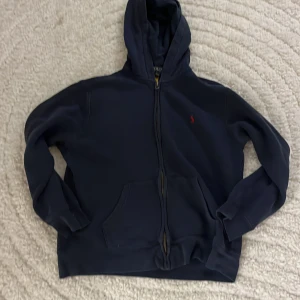 Mörkblå hoodie från Polo Ralph Lauren - Mörkblå hoodie från Polo Ralph Lauren med dragkedja och huva. Klassisk röd broderad logga på bröstet och stora fickor framtill. Skön och stilren modell i bomull, perfekt för chill dagar eller när du vill ha en clean look.