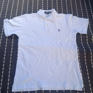 Vit pikétröja Polo Ralph Lauren M - Klassisk vit pikétröja från Polo Ralph lauren, Tröjan har korta ärmar, krage och knäppning framtill samt den ikoniska broderade loggan på bröstet. Tillverkad i bomull och passar perfekt till en clean och stilren look, stor i storleken 