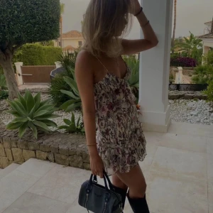 Blommig kort topp med volang - Supersöt kort topp med tunna axelband och volangdetalj. Toppen är ljus med blommigt mönster i lila, rosa och svart. V-ringad och med smock i midjan för en skön passform. Perfekt för varma dagar och sommarvibbar.