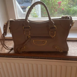 Beige handväska med guld detaljer - Säljer en beige handväska med guldiga metalldetaljer och dragkedja framtill. Väskan har både handtag samt dekorativa spännen och nitar. Dock har den små små rivmärken på undersidan av väskan, som man ser på sista bilden! Dom syns knappt!! Pris kan självklart diskuteras❤️