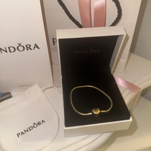 Guldarmband med hjärtlås från Pandora - Supersnyggt armband från Pandora i guldton med ett hjärtformat lås. Armbandet har en klassisk, stilren design och kommer i originalask med tillhörande påse och förpackning. Tror att det är 18cm, kan dubbelkolla vid intresse. Guldpläterad 