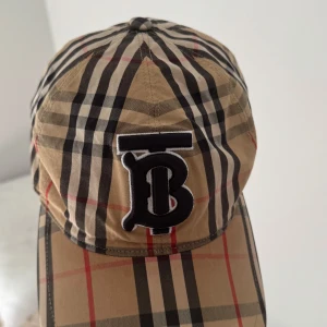 Burberry rutig keps med B-logga - Snygg keps från Burberry i klassiskt beige, svart och rött rutmönster. Stor broderad B-logga framtill. Tillverkad i 100% bomull och har den ikoniska Burberry-looken. Perfekt accessoar för att lyfta din outfit.