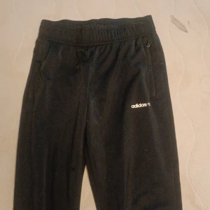 Svarta mjukisbyxor från Adidas - Svarta mjukisbyxor från Adidas med elastisk midja och dragkedjefickor. Byxorna har en smal passform och är tillverkade av återvunnen polyester, märkta med Adidas-logga på låret. Perfekta för chill eller träning.