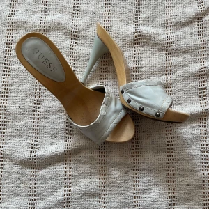 Guess ljusgrå klackade sandaler - Snygga ljusgrå sandaler från Guess med hög klack och träfärgad sula. Skorna har öppen tå, bred rem i skinn med dekorativa nitar och synliga sömmar. Perfekta för dig som vill sticka ut med en cool och trendig look.
