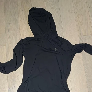 Svart tröja från Ralph Lauren - Svart hoodie från Ralph Lauren med klassisk logga broderad på bröstet. Tröjan är i mjuk bomull, har långa ärmar och en skön huva. Perfekt för en chill och stilren look. Tröjan är tunn