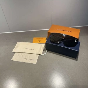 Svarta Louis Vuitton solglasögon - Snygga svarta solglasögon från Louis Vuitton med rektangulär form och LV-monogram på glasen. Bågarna är mattsvarta och tillverkade i ett lätt syntetmaterial. Kommer med originalbox, fodral och putsduk. Perfekt accessoar för en clean och lyxig look.