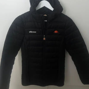  Jacka - Svart dunjacka från Ellesse med huva och dragkedja framtill. Jackan har quiltad design och logga på bröstet. Perfekt för kyliga dagar och enkel att matcha med olika outfits.