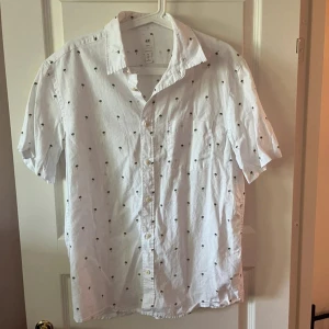 Vit kortärmad skjorta från H&M - Vit skjorta från H&M med korta ärmar och små svarta palmträd som mönster. Skjortan har klassisk krage, bröstficka och knäppning framtill. Perfekt för sommarens chill dagar och ger en avslappnad vibe.