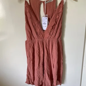 Rostrosa kort byxdress från Pull&Bear - Supersöt rostrosa kort byxdress från Pull&Bear med djup v-ringning och spetsdetaljer längs urringningen. Byxdressen är ärmlös och har en luftig, rynkad struktur i mjuk bomull. Perfekt för varma dagar och sommarhäng!