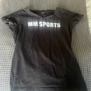 Svart t-shirt från MM Sports - Svart t-shirt från MM Sports med vit text på bröstet. Klassisk v-ringning och korta ärmar. Skön passform och tillverkad i mjukt bomullsmaterial, perfekt för träning eller chill. Enkel och stilren design som funkar till det mesta.