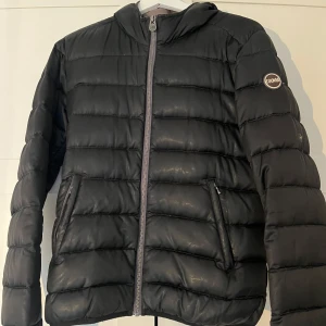 Svart dunjacka från Colmar - Säljer en svart dunjacka från Colmar med dragkedja och huva. Jackan har en quiltad design och två fickor med dragkedjor framtill (dock en trasig). Även en liten reva i fodret som tidigare varit lagad med en liten lapp. Italiensk storlek 46 motsvarar en S.