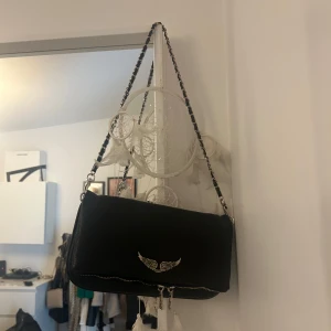 Zadig väska  - Snygg svart axelväska med kedjedetalj på axelremmen och ett coolt vingmotiv på framsidan. Perfekt för att ge din outfit en edgy touch. Väskan har en dragkedja för säker förvaring.