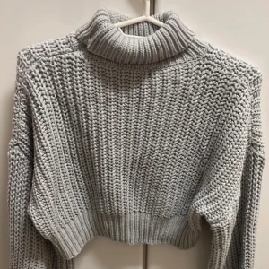 Grå stickad polotröja - Säljer en mysig grå stickad polotröja med en croppad design. Tröjan har en hög polokrage och långa ärmar, perfekt för kyliga dagar. Den är både stilren och bekväm, idealisk för höst och vinter.