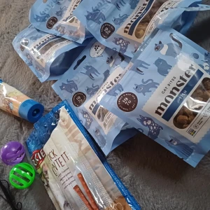 Monster Cat Bites - Säljer flera påsar med Monster Cat Bites, perfekta för att skämma bort din katt. Förpackningarna är ljusblå med söta kattillustrationer. Innehåller smakrika bitar som din katt kommer älska. Perfekt för kattägare som vill ge sina husdjur något extra gott. En påse öppnad. Säljer även katt sticks, en tagen och lever pastej på tub, öppnad med blivit klickad på tallrik. Även två bollar och en pinne