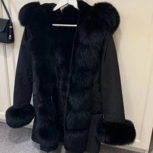 Svart pälsjacka med huva - Snygg svart jacka med fluffig pälsdetalj runt huva, ärmslut och framkant. Perfekt för att hålla sig varm och stilren under kyliga dagar. Köptes från hollies och pris kan diskuteras vid snabb affär! 