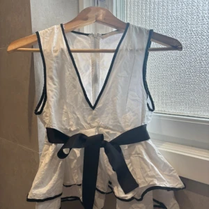 Vit ärmlös topp med svart knytband - Säljer en söt vit ärmlös topp med svart knytband i midjan och svarta detaljer. Toppen har en v-ringad design och dragkedja i ryggen. Perfekt för sommarens alla tillfällen! På andra bilden ser du att det har gått upp några trådar men inget som påverkar dragkedjan eller som man tänker på när den är på!