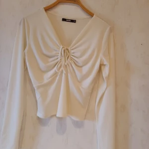 Vit långärmad topp från Bik Bok - Säljer en stilren vit långärmad topp från Bik Bok med v-ringning och snygg knytdetalj framtill. Perfekt för en chic och enkel look. Toppen är i ett mjukt och bekvämt material.