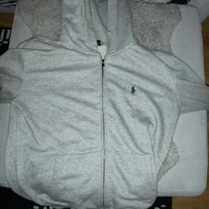 Grå hoodie från Ralph Lauren - Säljer en stilren grå hoodie från Ralph Lauren med dragkedja och det klassiska broderade logotypen på bröstet. Hoodien har en bekväm passform och är perfekt för en avslappnad look. Den är tillverkad i mjukt material och har en praktisk huva.