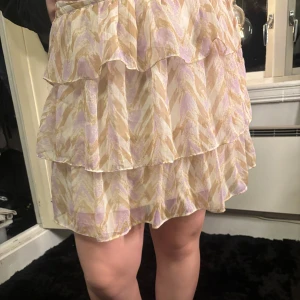 Mönstrad kjol från Vero Moda - Säljer en mönstrad kjol från Vero Moda i ljusa toner av beige och lila. Kjolen har volanger och ett elastiskt midjeband för en bekväm passform. Perfekt för en somrig look!