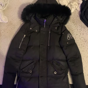  Moose Knuckles jacka - Säljer en svart dunjacka från Moose Knuckles med pälskantad huva och flera praktiska fickor med dragkedjor. Jackan har en stilren design med logotypdetaljer på ärmen och en quiltad insida för extra värme. Perfekt för kalla vinterdagar.