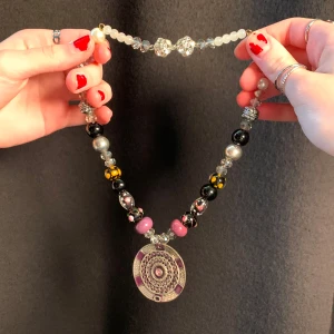 Färgglatt pärlhalsband med stort hänge - Unikt halsband med ett stort, runt hänge i silver och lila nyanser. Halsbandet har pärlor i olika färger och mönster, inklusive svarta, rosa och blommiga detaljer. Det finns inget lås till halsbandet eftersom det sitter fast via en gummitråd med knut. Halsbandet är i sin helhet ca 40 cm!
