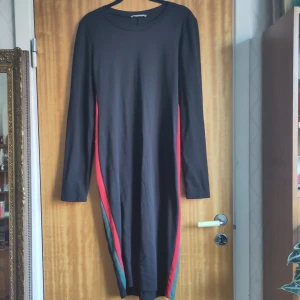 Svart klänning med färgglada ränder från Zara Trafaluc - Snygg svart långärmad klänning från Zara Trafaluc med färgglada ränder i rött och grönt längs sidorna. Perfekt för en stilren look med en twist. Klänningen har en enkel rund halsringning och är tillverkad i ett mjukt material.