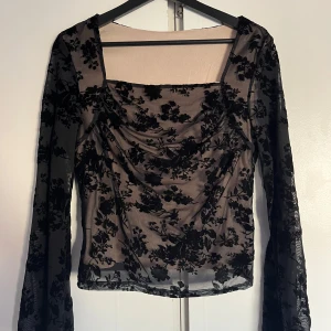 Svart blommig mesh-topp - Snygg svart mesh-topp med blommigt mönster. Toppen har långa ärmar och en draperad framsida som ger en elegant look. 