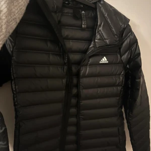 Svart dunjacka från Adidas - Säljer en snygg svart dunjacka från Adidas. Jackan har en quiltad design med dragkedja framtill och en praktisk huva. Perfekt för kyligare dagar och ger en sportig look. Strlk 38/40
