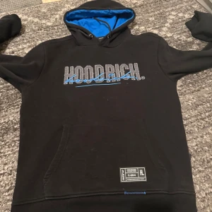 Svart hoodie från Hoodrich - Säljer en svart hoodie från Hoodrich med blå detaljer och tryck både fram och bak. Hoodien har en känguruficka och en justerbar huva med blått foder. Perfekt för en avslappnad stil.