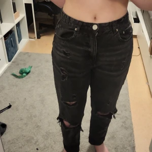 Svarta jeans från FB Sister - Säljer ett par svarta jeans från FB Sister i modellen 'Mom's Fit'. De har en cool slitningseffekt och är högmidjade med en knapp och dragkedja framtill. Perfekta för en avslappnad stil.