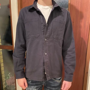 Svart overshirt med knappar - Snygg svart overshirt med knappar och två bröstfickor. Perfekt för lager-på-lager stil. Långärmad och tillverkad i ett robust material som ger en cool look.