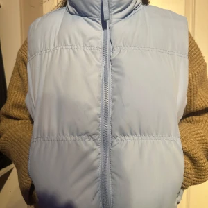Ljusblå dunväst med dragkedja - Säljer en ljusblå dunväst med dragkedja. Perfekt för kyliga dagar när du vill hålla värmen utan att bära en jacka. Västen har en hög krage och en stilren design.