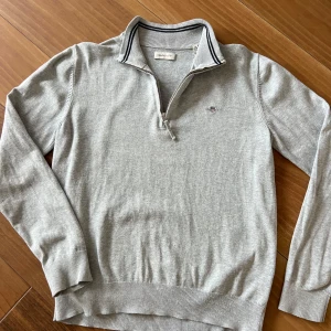 Grå halfzip från Gant - Säljer en stilren grå tröja från Gant med dragkedja och broderad logga på bröstet. Tröjan har långa ärmar och ribbade muddar vid ärmslut och nederkant. Perfekt för en avslappnad och snygg look.