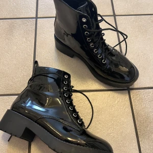 Svarta lackade boots med snörning - Snygga svarta boots i lackat material med snörning framtill. De har en robust sula med bra grepp och en klassisk design som passar perfekt för höst och vinter. Perfekta för att ge din outfit en edgy touch.