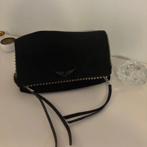 Zadig & voltaire nessiccär/ väska - Nypris: 4999kr Snygg svart axelväska i mocka med coola nitar längs kanterna. Väskan har en praktisk dragkedja och en justerbar axelrem. Perfekt för att ge din outfit en edgy touch.