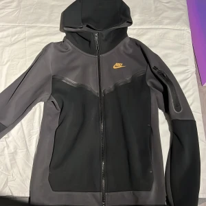 Svart och grå hoodie från Nike - Säljer en stilren svart och grå hoodie från Nike med dragkedja och huva nu när den har blivit för liten. Hoodien har en liten ficka med dragkedja på ärmen och Nike-loggan i gult på bröstet. 