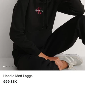 Svart hoodie med logga från Calvin Klein - Säljer en svart hoodie från Calvin Klein med en framträdande logga i rött på bröstet. Hoodien har en klassisk design med långa ärmar och en praktisk ficka framtill. Perfekt för en stilren och bekväm look.