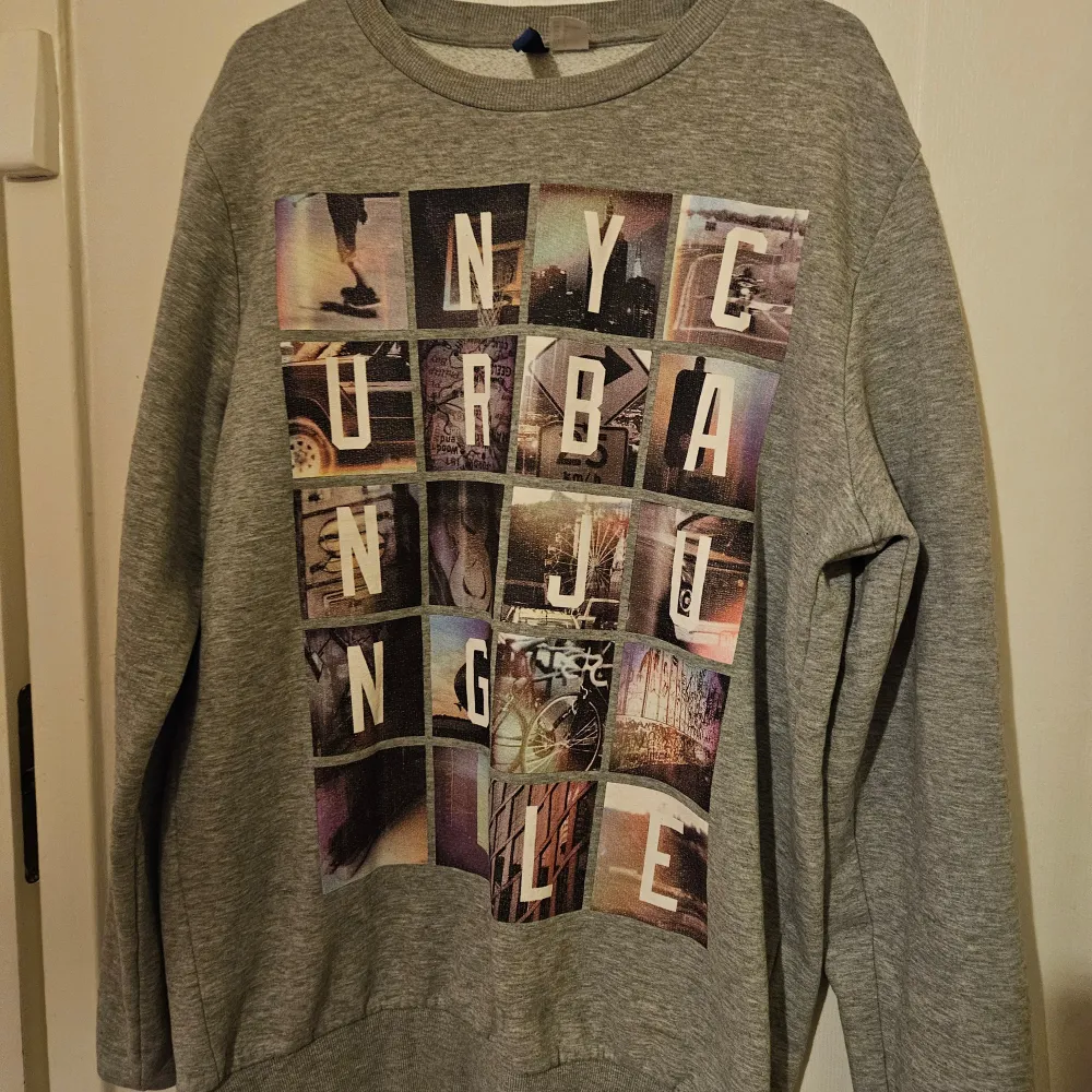 Säljer en grå sweatshirt från Divided med ett coolt tryck på framsidan som säger 'NYC Urban Jungle'. Tröjan har långa ärmar och är gjord av en mjuk bomullsblandning. Perfekt för en avslappnad stil.. Neuletakit & Villapaidat.