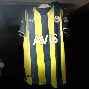 Fenerbache tröja från Puma - Snygg gul och blå randig fotbollströja från Puma med korta ärmar. Tröjan har sponsorloggor på bröstet och ärmarna, samt klubbemblem på framsidan. Perfekt för fotbollsfans som vill visa sitt stöd.