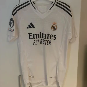 Vit Real Madrid fotbollströja - Säljer en vit Real Madrid fotbollströja från Adidas med korta ärmar. Tröjan har svarta detaljer och klubbens emblem på bröstet. På ryggen finns namnet 'Mbappé' och nummer 9. Perfekt för fans av laget!