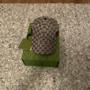 Mönstrad keps från Gucci - Snygg keps från Gucci med det klassiska GG-mönstret i beige och svart. Kepsen har en röd och grön randdetalj på sidan och justerbar passform. Perfekt för att ge din outfit en lyxig touch.