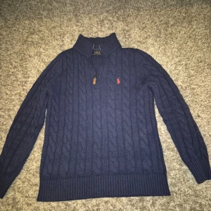 Mörkblå kabelstickad tröja från Polo Ralph Lauren - Säljer en snygg mörkblå kabelstickad tröja från Polo Ralph Lauren. Tröjan har en dragkedja vid halsen och ett broderat rött märke på bröstet. Perfekt för kyliga dagar med sin ribbade krage och muddar.