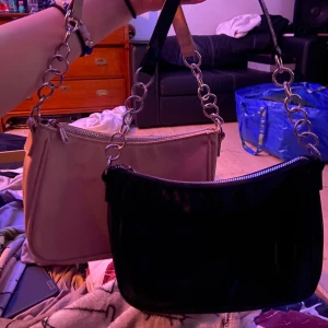 Svart och rosa axelväska med kedjedetaljer - Två stilrena axelväskor i svart och rosa med glänsande finish. Väskorna har kedjedetaljer på axelremmen och en praktisk dragkedja upptill. Perfekta för att ge en trendig touch till din outfit.