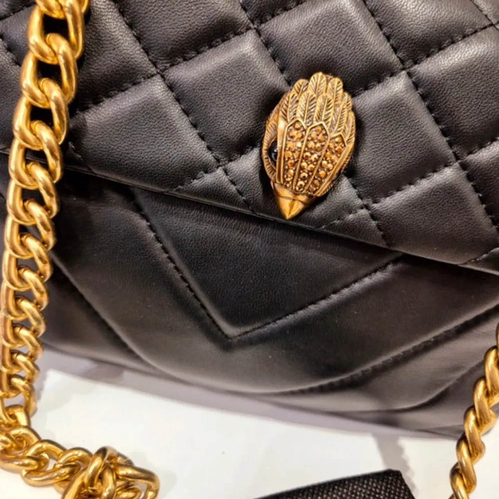 Snygg svart axelväska från Kurt Geiger med quiltad design och guldfärgad kedja. Väskan har en unik detalj på framsidan och är perfekt för att ge din outfit en lyxig touch. Den är tillverkad i skinn och har en elegant och tidlös stil.. Laukut & Käsilaukut.