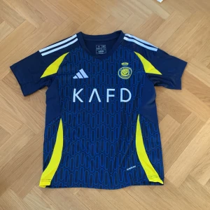 Al nassr borta ställ - Snygg blå fotbollströja från Adidas med gula detaljer och vit text med Ronaldo på ryggen. Tröjan är storlek small. Tröjan har korta ärmar och ett mönster över hela framsidan. Perfekt för fotbollsfans!