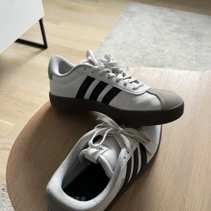 Vita Adidas sneakers med svarta ränder - Snygga vita Adidas sneakers med klassiska svarta ränder och brun sula. Perfekta för en avslappnad stil. Skorna har snörning och en stilren design.