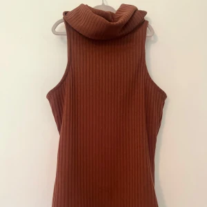 Brun ribbad ärmlös polotopp från River Island - Säljer en brun ribbad ärmlös polotopp från River Island i storlek 34. Toppen har hög krage och är tillverkad i ett mjukt, stretchigt material som sitter snyggt på kroppen. Perfekt att styla med jeans eller kjol för en trendig look.