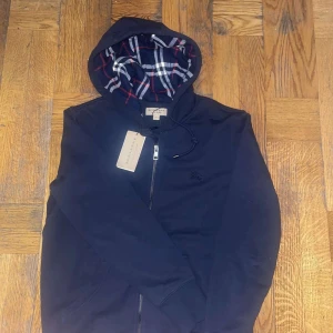 Mörkblå hoodie från Burberry - Snygg mörkblå hoodie från Burberry med dragkedja och klassiskt rutigt mönster i huvan. Perfekt för en stilren och bekväm look. Skicka prisförslag
